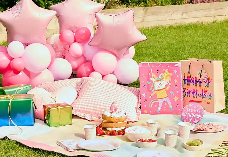 Pastel birthday picnic setup with pink balloons, gift bags, a birthday cake, party tableware, cushions, and a number 6 birthday decoration on a garden lawn.